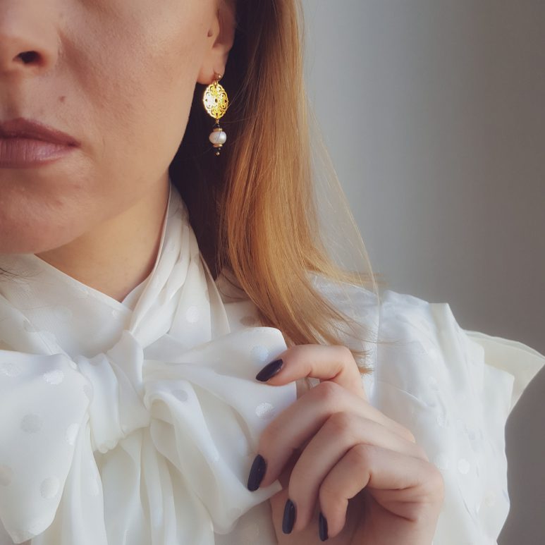 błyszczący kolczyk z perłą i złotą rozetą na modelce w białej koszuli, z wiązana pod szyją kokardą